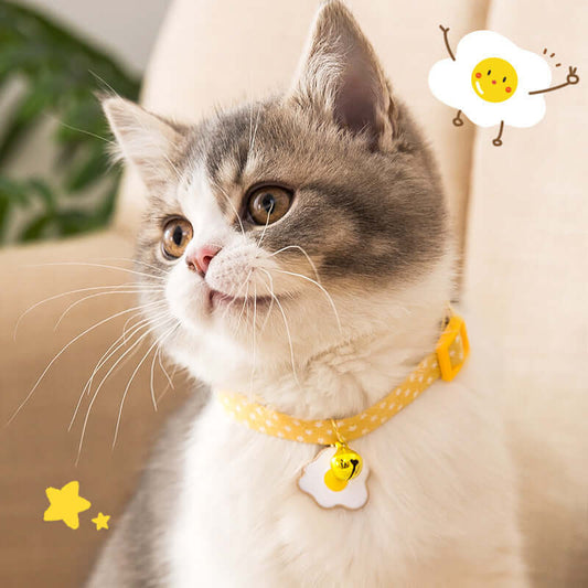 Cat wearing a yellow polka dot collar with a smiley face tag, sitting on a beige surface.