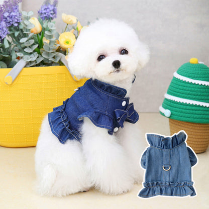 Small white dog wearing a blue denim dress with ruffles, standing next to a yellow basket with flowers and a green and white striped hat.